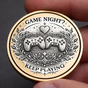 May include: A gold-colored coin with the words "GAME NIGHT?" and "KEEP PLAYING" encircling a design of two game controllers and a heart. The coin is held between fingers.