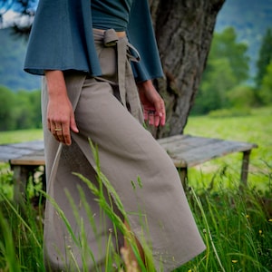 May include: A dark teal wrap top with wide, flared sleeves paired with high-waisted, light brown trousers featuring a side slit and a matching fabric tie belt at the waist. The trousers have a straight-leg cut and a clean, simple design.