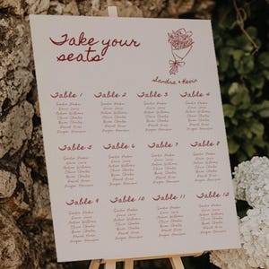 Könnte beinhalten: Ein weißes Sitzplan auf einer Holzstaffelei mit dem burgunderfarbenen Schriftzug "Take your seats". Der Plan listet Namen unter den Überschriften "Table 1" bis "Table 12" auf. Oben befindet sich eine Blumenillustration. Weiße Blumen und eine Steinmauer sind im Hintergrund.