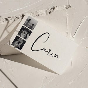 Könnte beinhalten: Eine weiße Karte mit dem Namen "Carin" in eleganter schwarzer Schrift. Die Karte zeigt drei kleine Schwarz-Weiß-Fotos. Die Karte liegt auf einer strukturierten weißen Oberfläche, mit einem getrockneten Blumenstiel im Hintergrund.