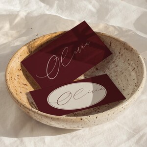 Könnte beinhalten: Zwei weinrote Visitenkarten mit dem Namen "Olivia" in eleganter weißer Schrift werden in einer gesprenkelten Keramikschale präsentiert. Eine Karte hat ein weißes Oval mit dem Namen. Die Schale steht auf einer weißen, strukturierten Oberfläche.