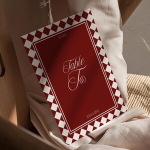 May include: A red and white table card with the words "Table Two" in elegant script. The card has a diamond pattern border and is set on a cream-colored fabric, suggesting a wedding or event theme.