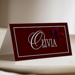 May include: A burgundy place card with the name "Olivia" in elegant white script. Two dark red roses are in the upper right corner. The card has a white border and is folded in half, standing on a white plate.