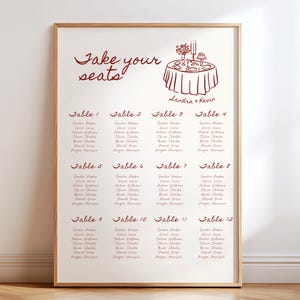 Könnte beinhalten: Ein gerahmter Hochzeits-Sitzplan mit dem burgunderfarbenen Schriftzug "Take your seats". Die Tabelle listet Tischnummern und Gästenamen auf, zusammen mit einer kleinen Illustration einer Tischdekoration. Der Rahmen hat eine helle Holzfarbe.
