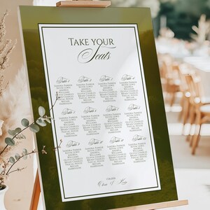 May include: A wedding seating chart on an easel. The chart has a green border and a white interior with the words "Take Your Seats" in elegant script. Names are listed under table numbers. The easel is made of wood.