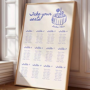 Könnte beinhalten: Ein gerahmter Hochzeits-Sitzplan mit hellbeigem Hintergrund und blauer Schrift. Oben steht "Take your seats" und eine Tischillustration. Die Namen sind nach Tischnummern geordnet.