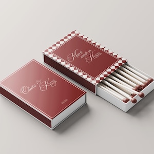Peut inclure: Deux boîtes d'allumettes rouges avec des bordures blanches. L'une est fermée, avec les noms "Olivia & Kevin" en écriture cursive. L'autre est ouverte, révélant des allumettes à têtes marron. Le dessus de la boîte ouverte indique "Match made in Heaven".