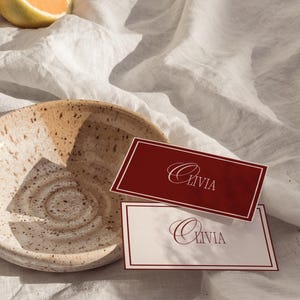 Könnte beinhalten: Eine gesprenkelte Keramikschale mit dunklem Rand liegt auf einem weißen Leinentuch. Zwei rechteckige Tischkarten mit dem Namen "Olivia" sind abgebildet. Eine Karte ist weinrot mit weißem Text, die andere ist weiß mit weinrotem Text.