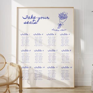 Könnte beinhalten: Ein gerahmter Hochzeits-Sitzplan mit blauem Text auf cremefarbenem Hintergrund. Der Text enthält "Take your seats" und Gästenamen unter den Tischnummern. Eine Blumenillustration ist ebenfalls vorhanden.