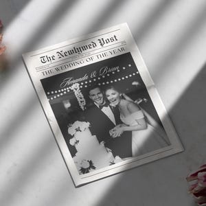 May include: A black and white newspaper titled "The Newlywed Post" featuring a wedding photo of a smiling couple, Amanda & Brian. The headline reads "The Wedding of the Year". The newspaper is on a white surface with a shadow.