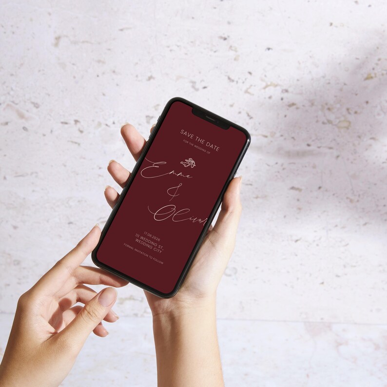 May include: A smartphone displays a burgundy-colored wedding invitation with the text "Save the Date." The invitation features elegant cursive names and floral accents. The phone is held in two hands.