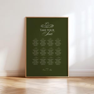 May include: A framed seating chart with a dark green background. The chart features elegant white text, including the words "Take Your Seats" and guest names. Two swans are at the top. The frame is gold.