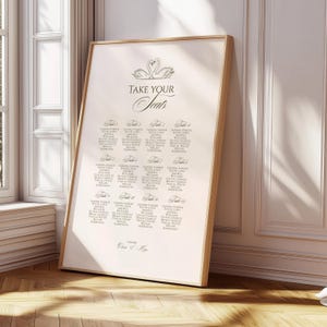 May include: A framed seating chart with a light beige background and a wooden frame. The text "TAKE YOUR SEATS" is at the top, with a swan illustration. Names are listed below, organized by table number. The date is printed at the bottom.