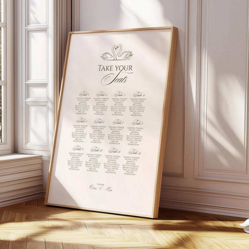 May include: A framed seating chart with a light beige background and a wooden frame. The text "TAKE YOUR SEATS" is at the top, with a swan illustration. Names are listed below, organized by table number. The date is printed at the bottom.
