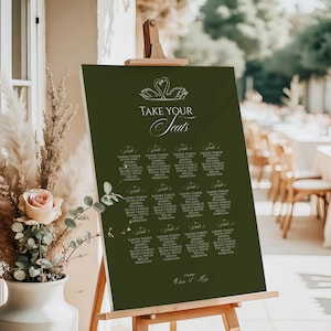May include: A dark green seating chart on a wooden easel. The chart features elegant white text that reads "TAKE YOUR Seats" above a list of names. Two swan illustrations are at the top. The date is written at the bottom.