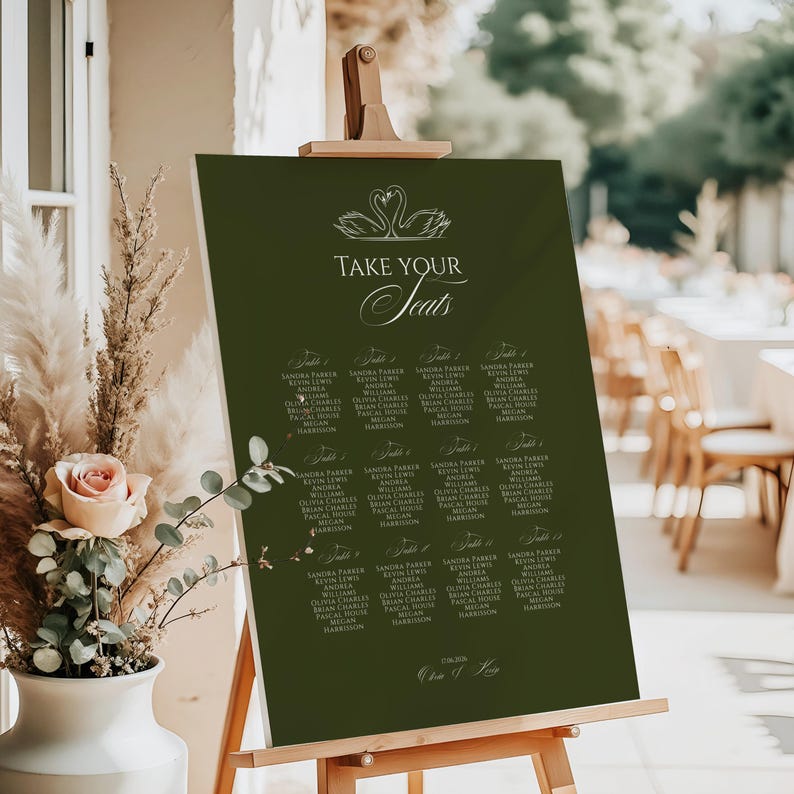 May include: A dark green seating chart on a wooden easel. The chart features elegant white text that reads "TAKE YOUR Seats" above a list of names. Two swan illustrations are at the top. The date is written at the bottom.