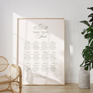 May include: A large, framed seating chart with the words "TAKE YOUR Seats" in elegant script. The chart lists names under table numbers, with a swan illustration at the top. The overall color scheme is neutral, with a light background and dark text.