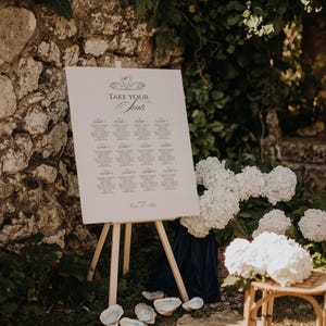 May include: A white sign on a wooden easel with the words "Take Your Seats" in elegant script, listing names and table assignments. The sign is decorated with a swan design. The scene is set outdoors with white flowers and a stone wall.
