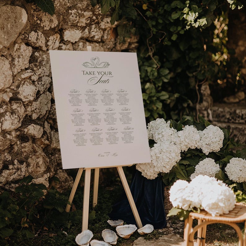 May include: A white sign on a wooden easel with the words "Take Your Seats" in elegant script, listing names and table assignments. The sign is decorated with a swan design. The scene is set outdoors with white flowers and a stone wall.