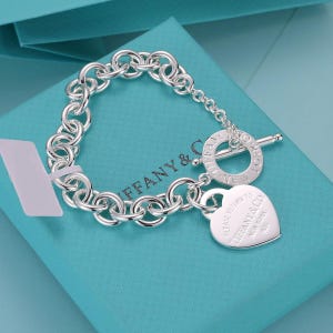Op de afbeelding: Een schakelarmband van sterling zilver met een knevelsluiting en een hartvormige hanger. De sluitring is gegraveerd met Tiffany & Co. New York en het hartvormige plaatje draagt de tekst Please Return To Tiffany & Co. New York 925.