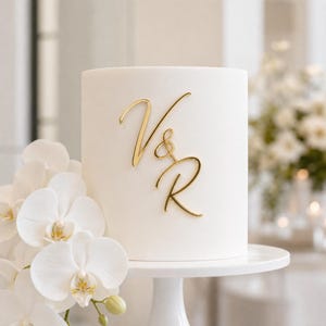 May include: A cylindrical white cake featuring a gold-colored script monogram of the letters V and R with an ampersand symbol. The cake is displayed on a white pedestal stand, positioned next to a cluster of white orchid flowers.