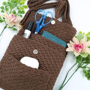 May include: A brown crochet crossbody bag with a long strap and tassel. The bag's open flap reveals a pair of blue scissors, a black marker, and a teal item. A white earbud case is in the front pocket. Artificial flowers are in the background.