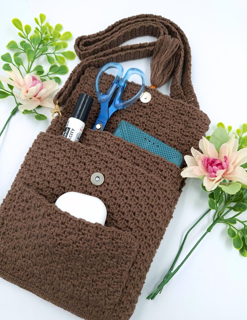 May include: A brown crochet crossbody bag with a long strap and tassel. The bag's open flap reveals a pair of blue scissors, a black marker, and a teal item. A white earbud case is in the front pocket. Artificial flowers are in the background.