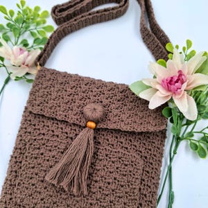 May include: A brown crochet crossbody bag with a flap closure and a long strap. The bag features a decorative tassel with a wooden bead. The bag is displayed with pink and green floral accents.