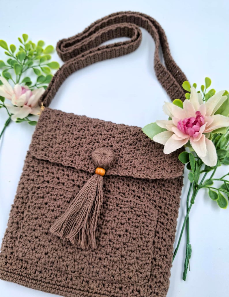 May include: A brown crochet crossbody bag with a flap closure and a long strap. The bag features a decorative tassel with a wooden bead. The bag is displayed with pink and green floral accents.