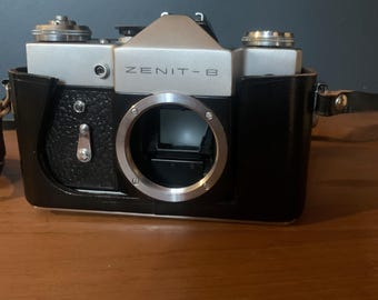 Puede incluir: Una cámara de película Zenit-B de 35 mm plateada y negra vintage con una funda de cuero negra. La cámara tiene una montura de lente prominente y varios diales y perillas. El texto "ZENIT-B" está impreso en la parte frontal de la cámara.