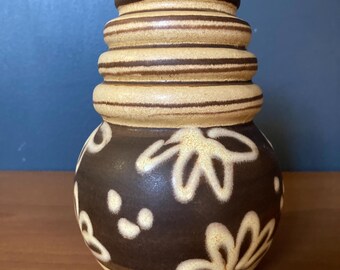 May include: A brown ceramic vase with a bulbous base and a stacked, cylindrical neck. The base is decorated with white floral and dot patterns. The neck has horizontal brown stripes. The vase sits on a wooden surface.