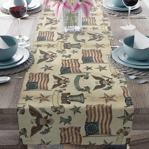 May include: A table runner featuring a vintage-style print of American flags, bald eagles, liberty bells, and stars on a cream background. The design includes patriotic motifs arranged in a repeating pattern across the fabric surface.