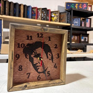 Puede incluir: Un reloj cuadrado de madera con una esfera marrón y un marco de madera marrón oscuro. La esfera del reloj presenta una silueta de un rostro. Las manecillas del reloj son negras con un segundero rojo. Los números están en una fuente clásica.