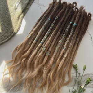 May include: A set of synthetic dreadlock extensions featuring a gradient color transition from dark brown at the roots to light blonde at the wavy ends. The dreadlocks are decorated with light blue thread wraps and small silver-toned metal charms.