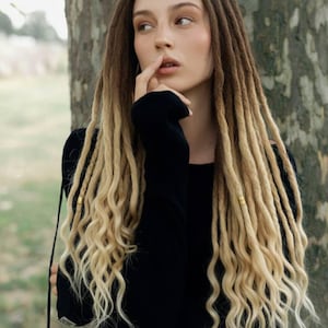 May include: A person with long, two-toned dreadlocks, transitioning from dark brown to blonde. The dreadlocks are styled with loose, wavy ends. The person is wearing a black long-sleeved top.
