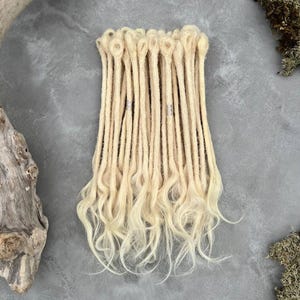 May include: A set of light blonde synthetic dreadlocks featuring a looped top for attachment. The strands are textured and cylindrical, transitioning into loose, wavy ends. Two of the dreadlocks are adorned with small, metallic silver beads.