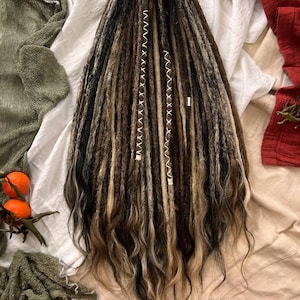 Puede incluir: Un conjunto de rastas sintéticas en tonos marrón, negro y rubio. Las rastas están dispuestas en cascada, con una mezcla de texturas y colores. Dos mechones están adornados con hilo blanco. El cabello está peinado sobre una tela blanca.