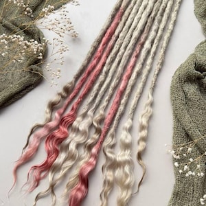Puede incluir: Un conjunto de extensiones de rastas sintéticas con textura ondulada. Los mechones son principalmente de color rubio claro o blanco roto, con varias rastas individuales que incorporan secciones de tonos rosa suave y rosa apagado a lo largo.