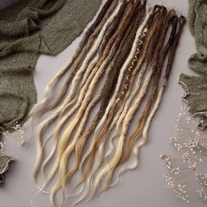 Peut inclure: Un ensemble d'extensions de dreadlocks synthétiques présentant une palette de couleurs dégradées allant du brun foncé aux racines au blond clair aux pointes. Les mèches ont un aspect texturé et mat avec des extrémités ondulées.