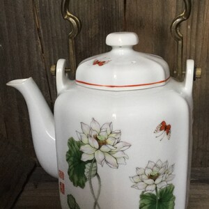 May include: White porcelain teapot with a brass handle and lid. The teapot is decorated with a floral design of white lotuses and green leaves, and red butterflies.