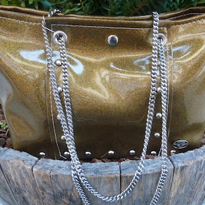 May include: A gold glitter tote bag with silver chain straps and silver studs. The bag is sitting on a wooden surface.