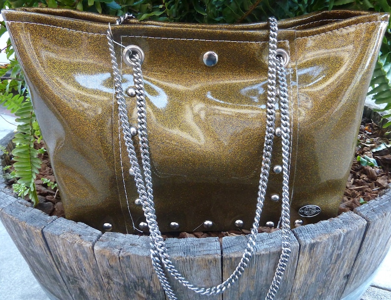 May include: A gold glitter tote bag with silver chain straps and silver studs. The bag is sitting on a wooden surface.