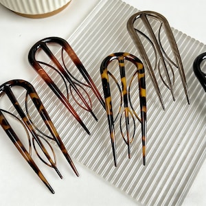 Puede incluir: Una colección de horquillas para el cabello en varios colores y patrones. Las horquillas tienen forma de U con recortes decorativos. Los colores incluyen marrón, negro y patrones de carey. Las horquillas se muestran sobre una superficie transparente y texturizada.