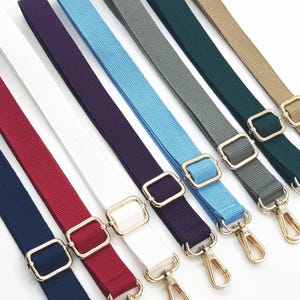 Puede incluir: Variedad de correas de bolso en azul marino, burdeos, blanco, morado, azul claro, gris, verde oscuro y beige. Cada correa tiene herrajes metálicos dorados, incluyendo hebillas rectangulares y mosquetones giratorios.