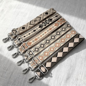 Może przedstawiać: Sześć pasków do toreb z wzorami i srebrnymi metalowymi elementami. Paski mają geometryczne wzory w różnych kolorach, w tym beżowym, czarnym, brązowym i szarym. Każdy pasek ma metalowe zapięcie na obu końcach.