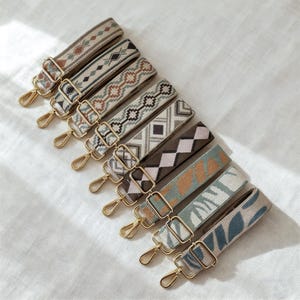 May include: Eight patterned bag straps in a variety of designs and colours. The straps feature gold-coloured metal clasps and buckles. The patterns include geometric and floral motifs in shades of brown, beige, teal, and grey. The straps are arranged diagonally.