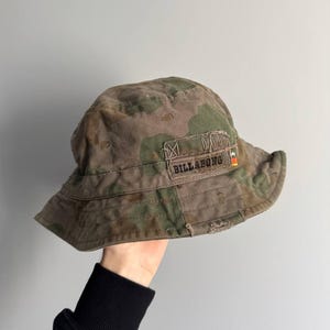 Puede incluir: Un sombrero de pescador de camuflaje con el nombre de la marca "BILLABONG" en un parche. El sombrero presenta un aspecto desgastado y envejecido con una mezcla de colores verdes, marrones y beige. El sombrero se sostiene sobre un fondo gris liso.