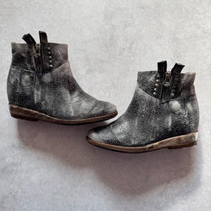 May include: A pair of ankle boots featuring a distressed black and white textured finish, side zippers, and small metallic stud accents along the collar. The boots have a low heel and a rounded toe design with a weathered appearance throughout.