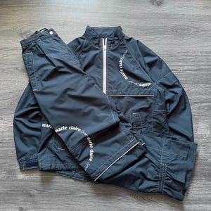May include: A navy blue tracksuit set, including a jacket and pants, with white trim and the text "marie claire sport". The jacket has a pink zipper and the pants have an elastic waistband.