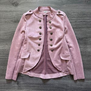 Peut inclure: Veste rose poudré de style militaire. Elle présente un devant croisé avec des boutons décoratifs, des épaulettes et deux poches avant. Le tissu a une texture douce semblable à du daim. Vêtement élégant.